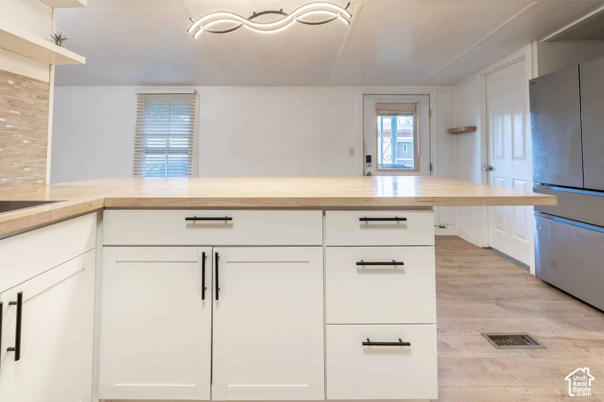 White shaker-style cabinetry with black hardware, extended wood-finish countertop, stainless steel refrigerator, and wood-finish flooring