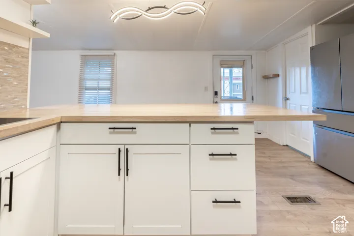 White shaker-style cabinetry with black hardware, extended wood-finish countertop, stainless steel refrigerator, and wood-finish flooring