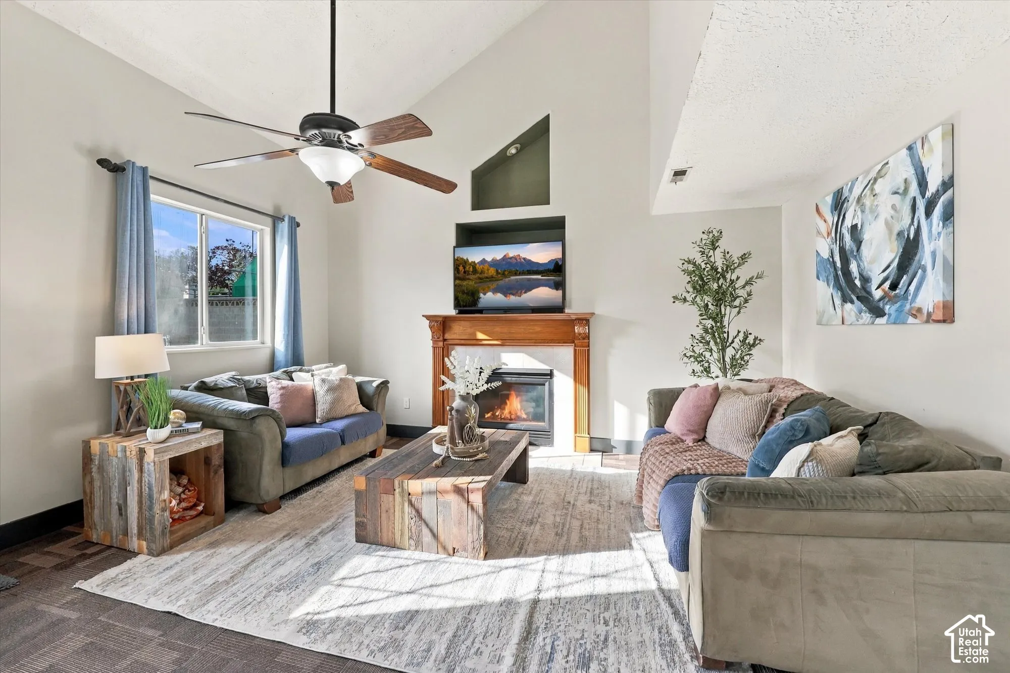 Vaulted ceiling with a ceiling fan, fireplace with a wood-finish mantle, window with blue drapery, and carpet.