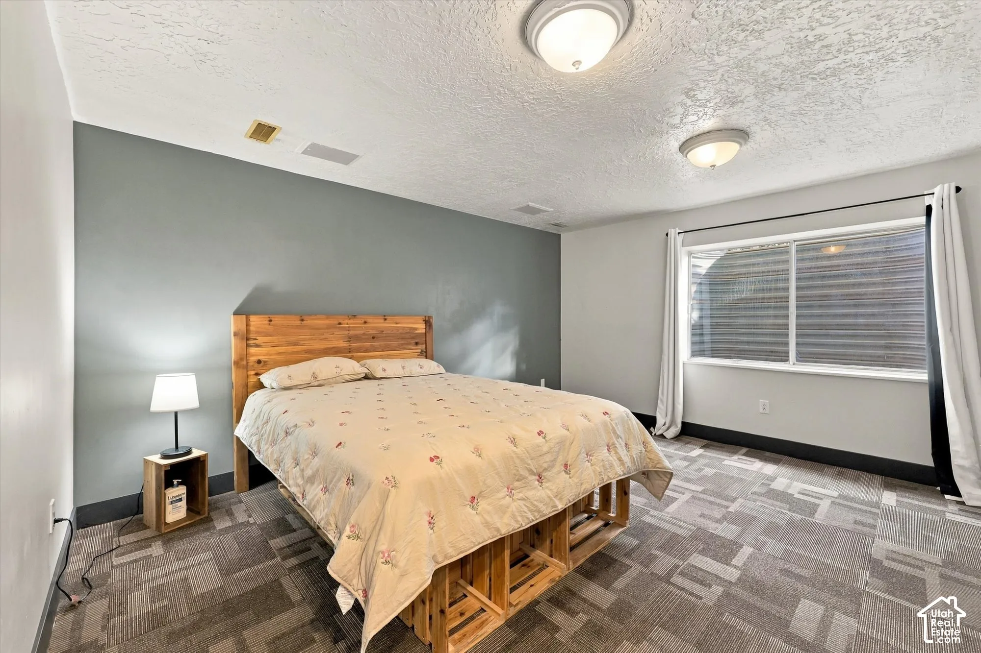 Window with blinds, wood-finish headboard, geometric pattern carpeting, two flush mount ceiling lights, and two-tone wall paint