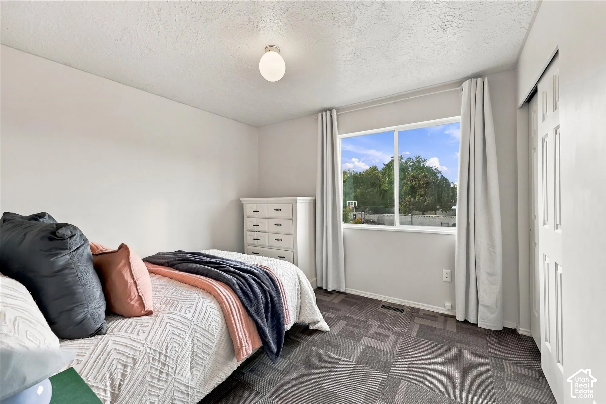 Bright secondary bedroom featuring large windows with natural light, neutral wall finishes, gray patterned carpet.
