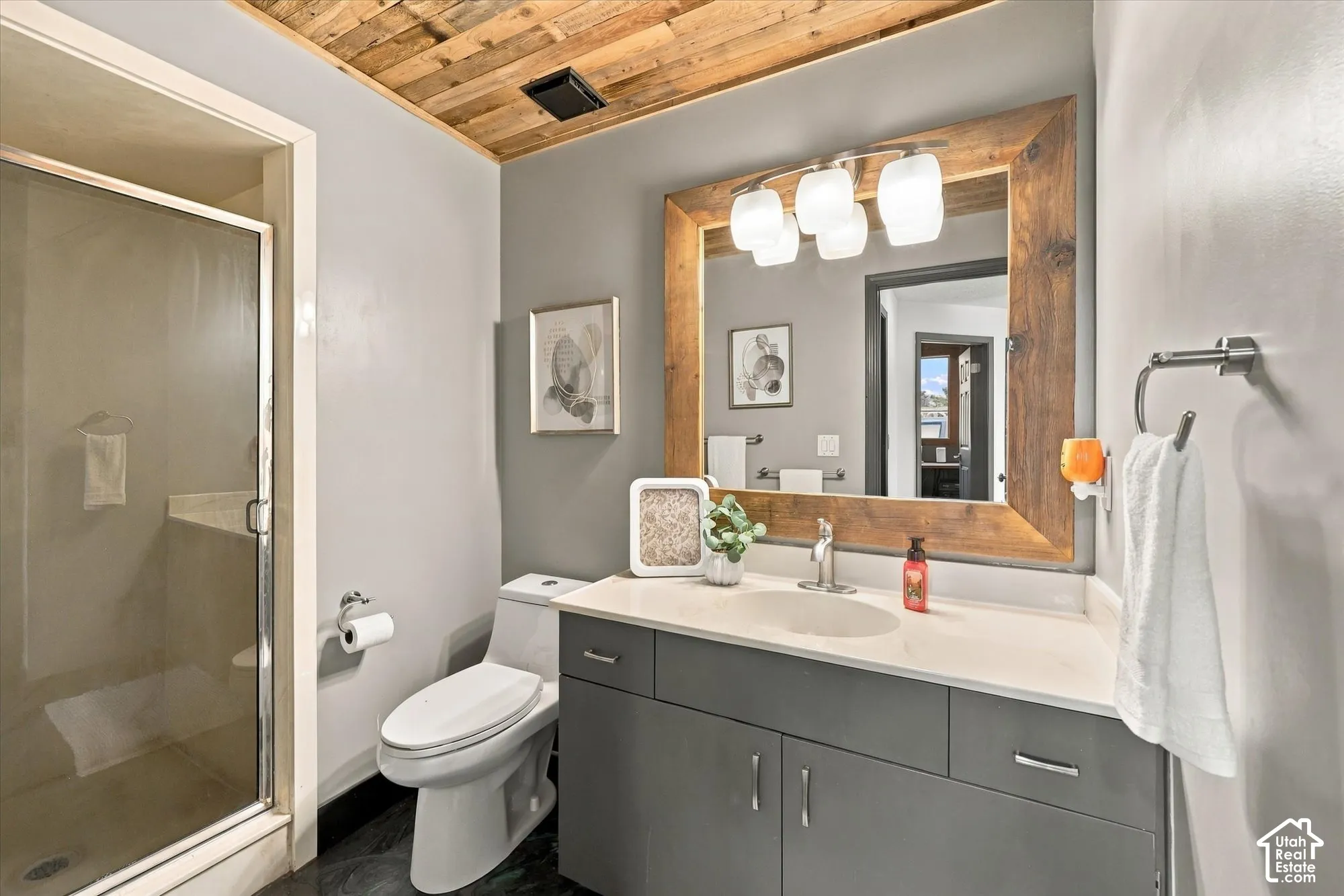 Wood-paneled ceiling, dark blue gray vanity with chrome hardware, wood-framed mirror with multi-bulb vanity light, and a glass-enclosed shower.