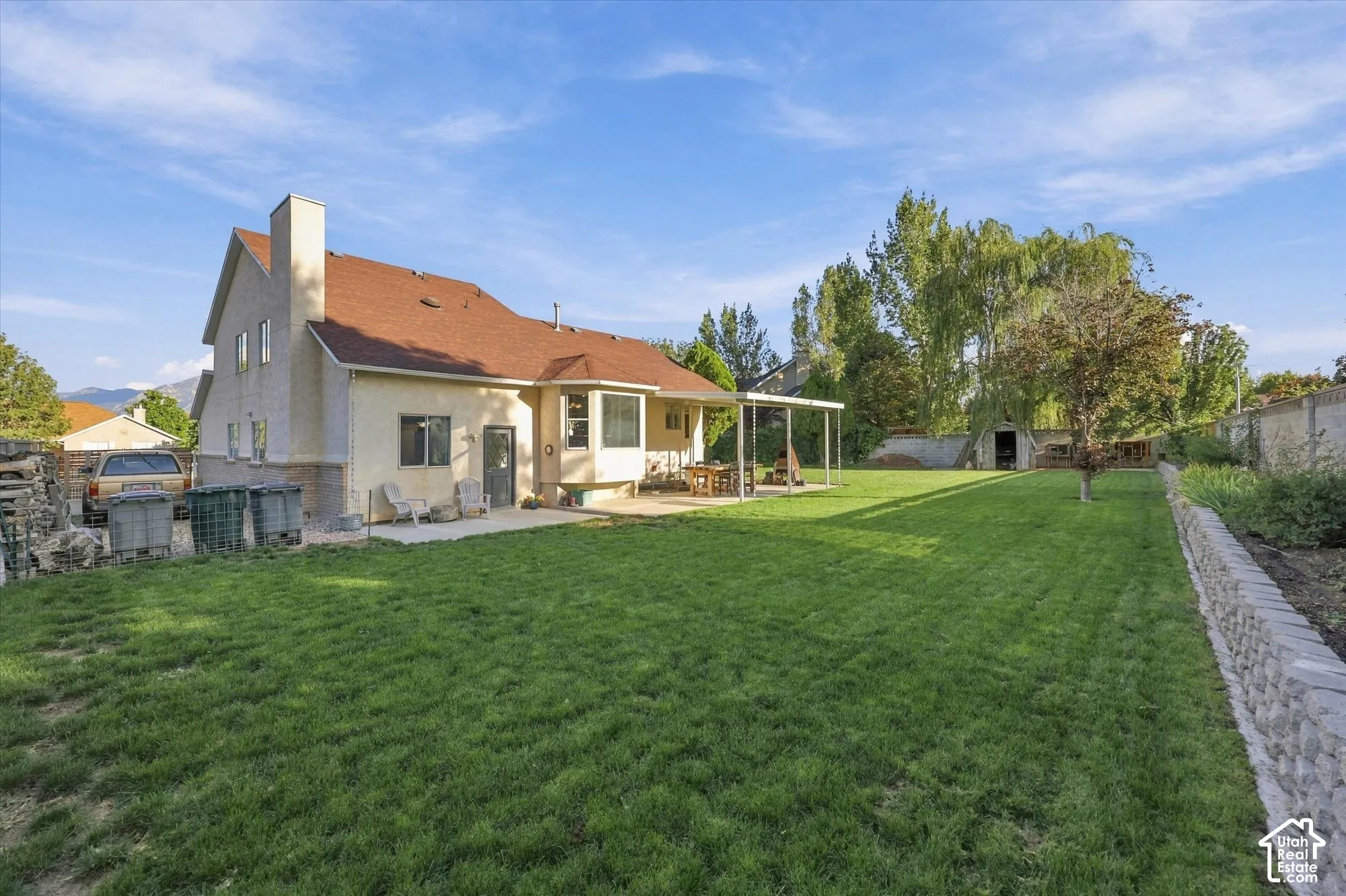 Expansive grassy yard with a covered patio, stone retaining wall, and an exterior storage shed