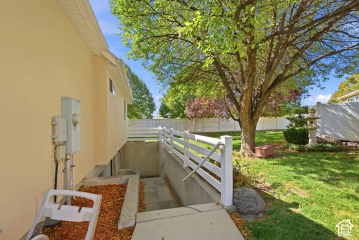 Exterior stucco finish, basement entrance with concrete steps, white picket-style railing, established lawn, and mature trees