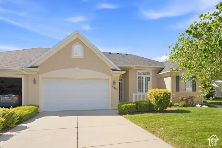 Stucco exterior with a two-car garage, pitched roof, front entry with transom window, and mature landscaping