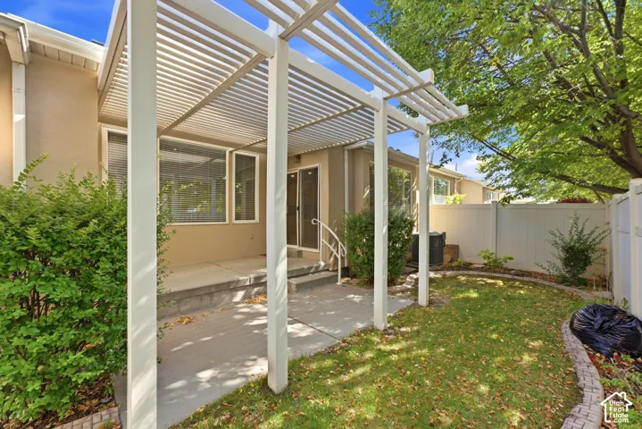 Patio featuring a white slatted pergola, concrete surface, sliding glass door, and adjacent lawn area with mature landscaping