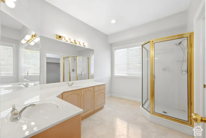 Dual vanity with integrated sinks, expansive mirror, wood-finish cabinetry, corner shower with brass-finish frame, and tile flooring