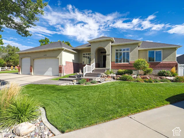 Stucco exterior with brick accents, two-car garage, concrete driveway, front lawn, and landscaped garden beds