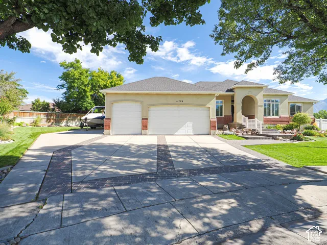 Stucco exterior with brick accents, arched entry, two-car garage, mature landscaping, and an expansive concrete driveway with brick inlay