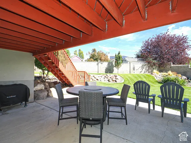Covered concrete patio with direct access to a terraced backyard, featuring a manicured lawn, established landscaping, and a wooden exterior staircase