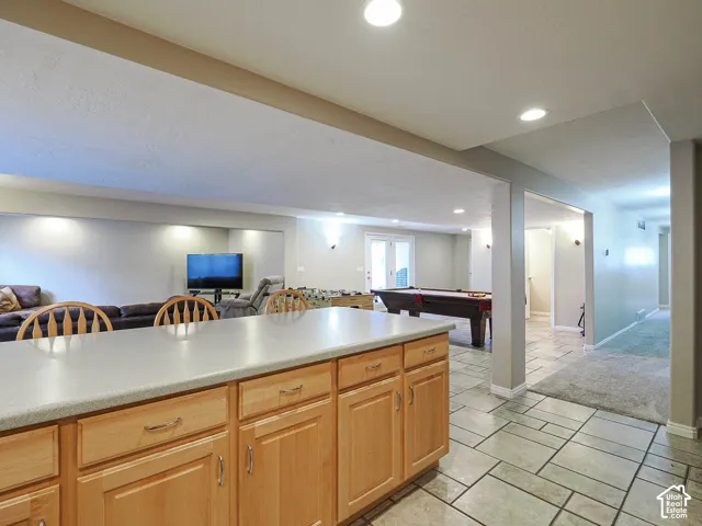 Spacious lower-level area featuring a large island with light wood cabinetry and a light-toned countertop, recessed lighting, and a combination of tile and carpet flooring