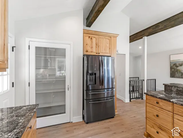 Vaulted ceiling with exposed wood beams, stainless steel refrigerator, wood-finish flooring, glass-paneled pantry door, and natural wood cabinetry with stone countertops