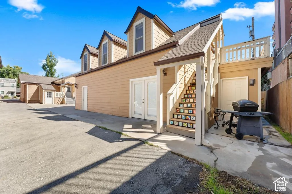 Multiple dormer windows, exterior staircase with decorative risers, upper-level deck with white railing, paved courtyard, and a detached building
