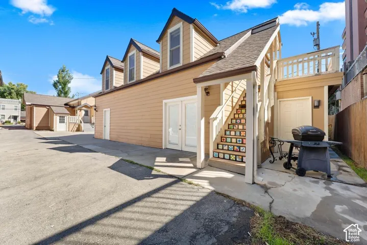 Multiple dormer windows, exterior staircase with decorative risers, upper-level deck with white railing, paved courtyard, and a detached building