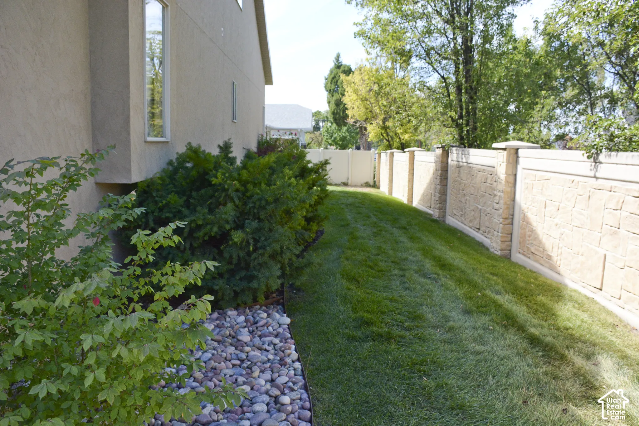 Stucco exterior featuring a bay window, natural landscaping with river rock, and a manicured lawn bordered by a stone and stucco privacy wall with decorative pilasters