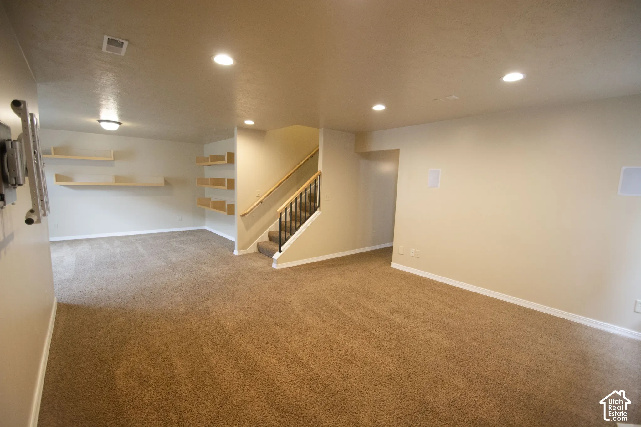 Spacious carpeted room featuring recessed lighting, built-in wall speakers, a staircase with a wooden handrail and wrought iron balusters, and integrated shelving