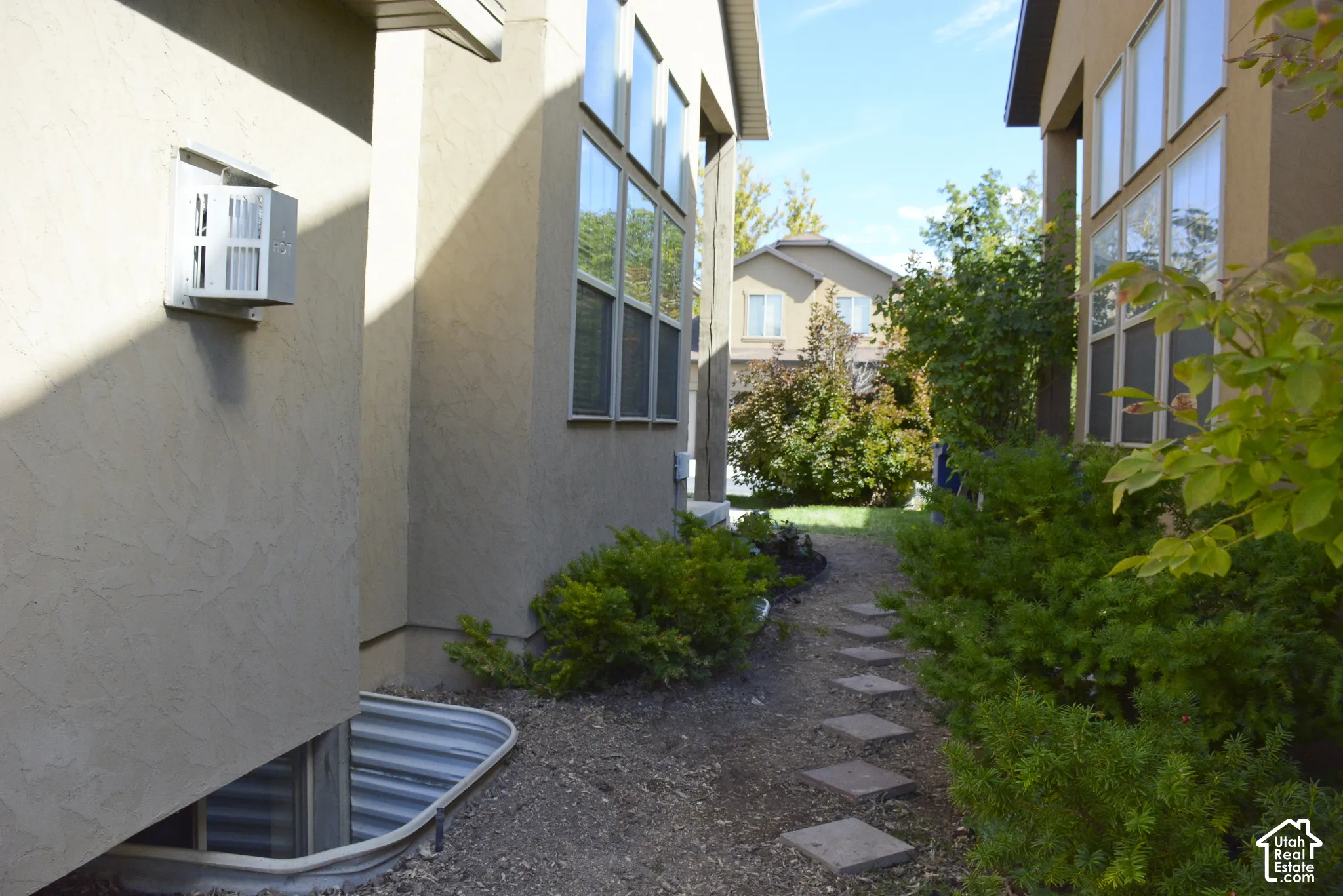 Stucco exterior with multiple windows, leading to a landscaped pathway with square stepping stones and mature shrubs