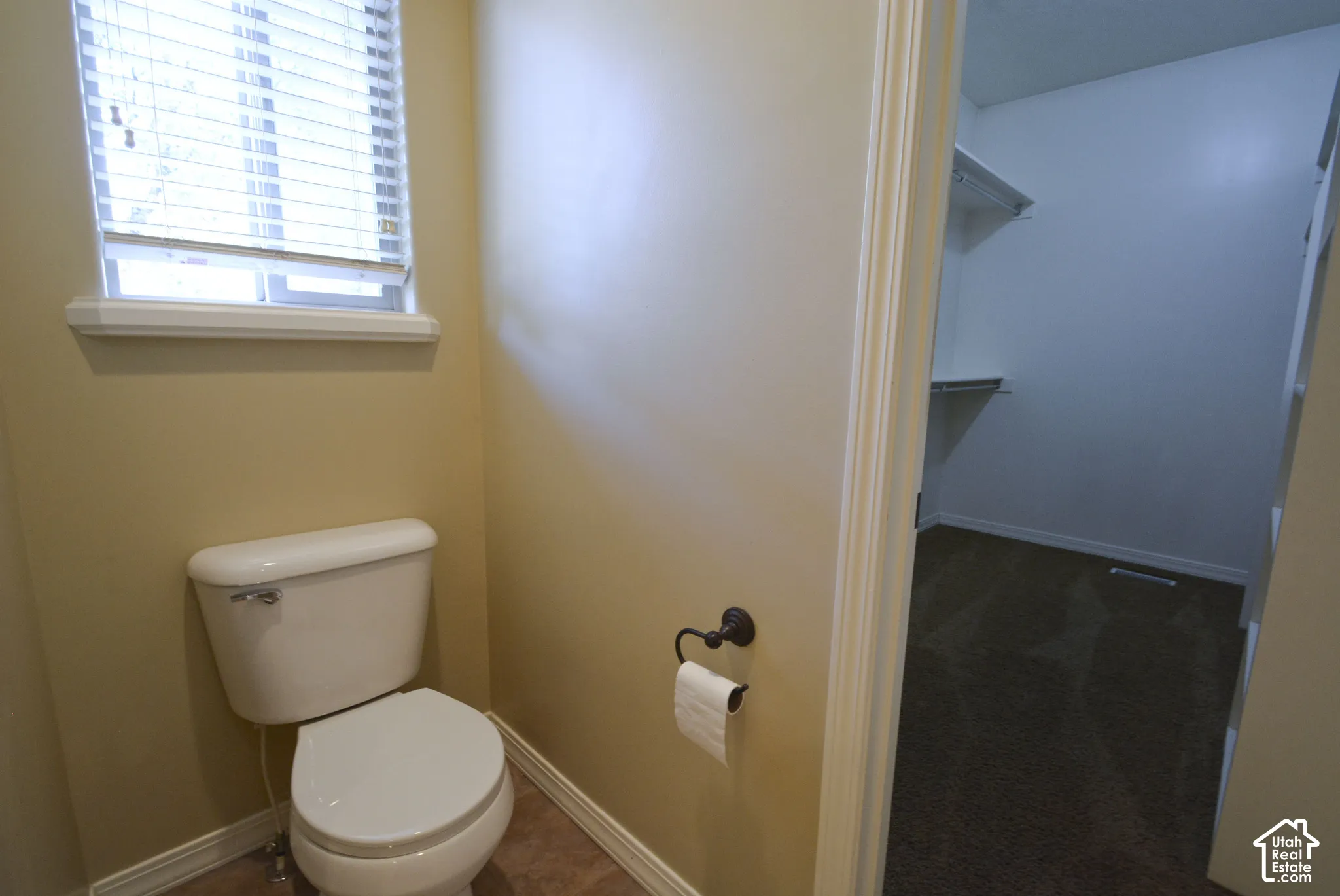 White toilet with a window, tile flooring, and an adjacent walk-in closet with carpet flooring and shelving