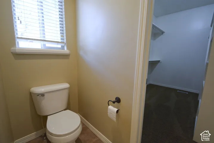 White toilet with a window, tile flooring, and an adjacent walk-in closet with carpet flooring and shelving