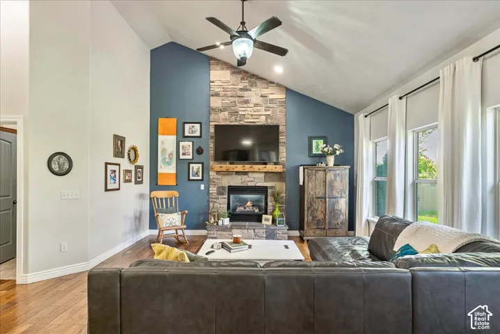 Vaulted ceiling with a ceiling fan, stone-clad fireplace with a wood mantle, wood-finish flooring, and multiple windows with roller shades