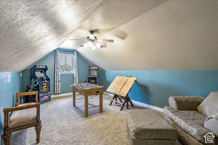 Sloped ceiling with textured finish, carpeted flooring, window with trim, ceiling fan, and painted walls
