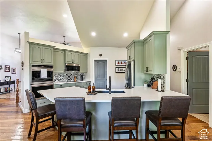 Kitchen featuring sage green cabinetry, stainless steel double wall ovens, a patterned tile backsplash, a large kitchen island with counter seating, and wood-finish flooring