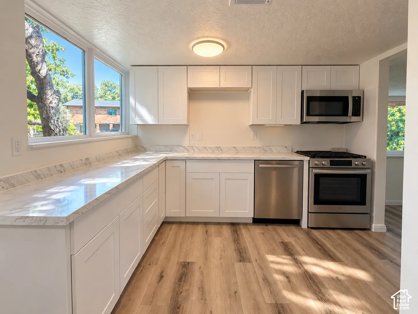 Kitchen featuring white shaker cabinetry, light-toned wood-finish flooring, stainless steel appliances, marble-look countertops, and ample natural light