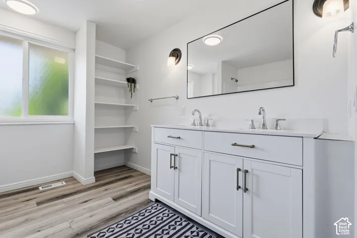 Dual vanity with integrated sinks, white cabinetry with dark hardware, a large wall-mounted mirror, and wood-finish flooring