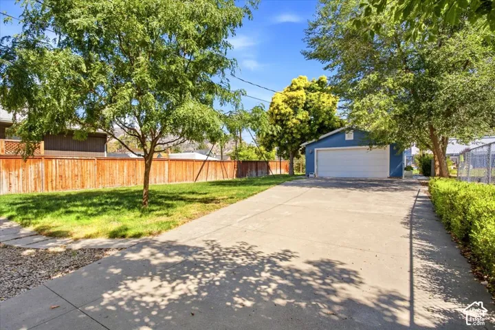 Detached blue structure featuring a white garage door, concrete driveway, manicured lawn, and mature trees