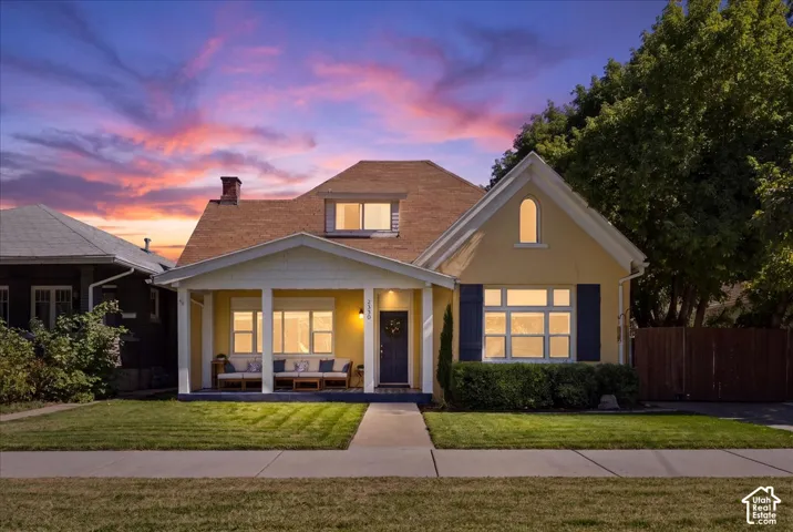 Yellow exterior with white trim, covered front porch, front entry door, front yard with manicured lawn, and mature landscaping