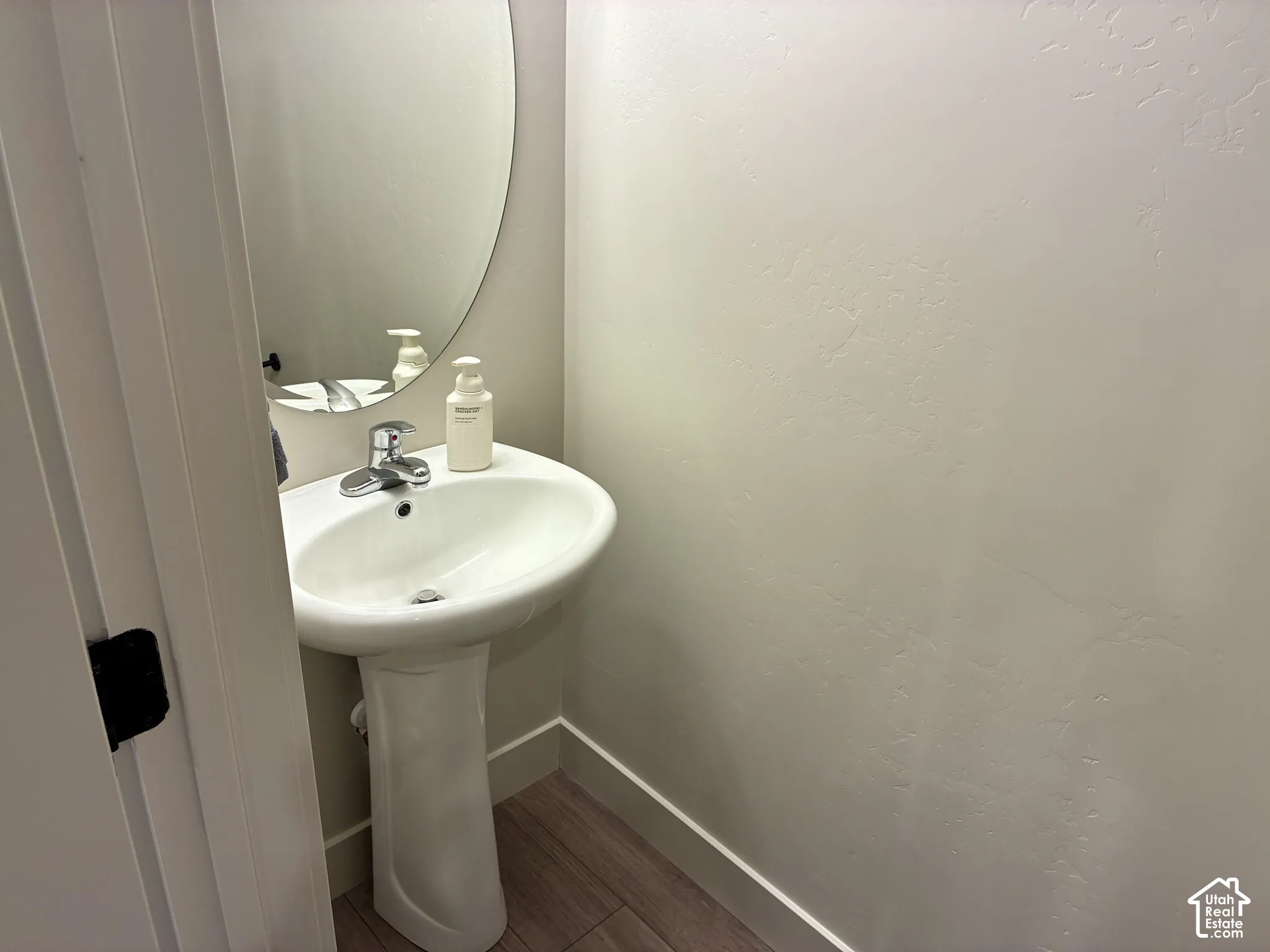 Half-bathroom adjacent to Kitchen and Garage entry. Pedestal sink with chrome faucet, oval mirror, and wood-finish flooring