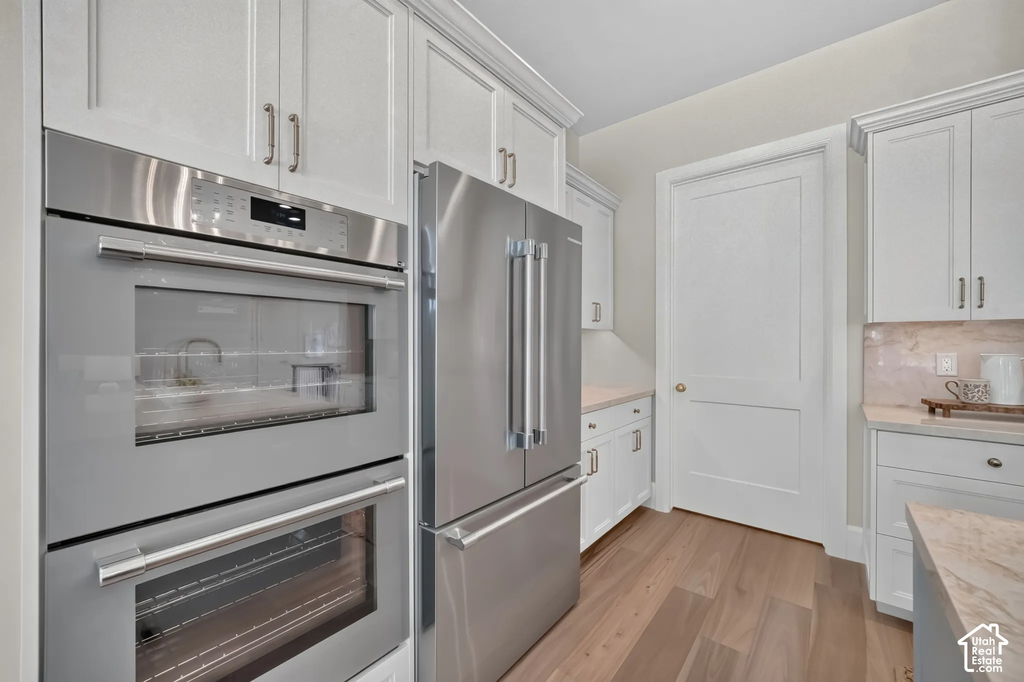 Built-in stainless steel double ovens, stainless steel refrigerator, white shaker-style cabinetry, light wood-finish flooring, and light-toned countertops