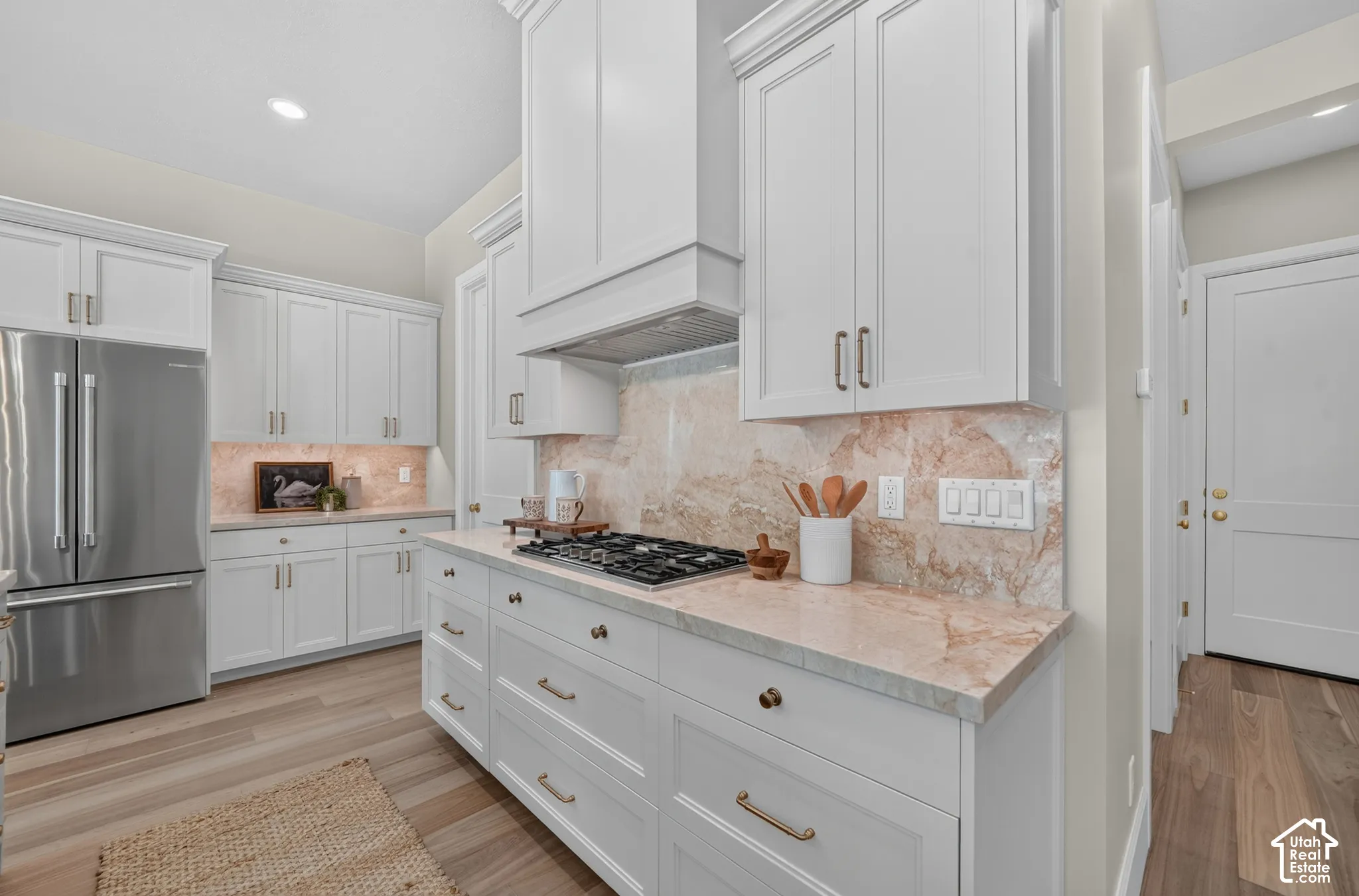White shaker cabinetry with brass-finish hardware, light-toned stone countertops, a coordinating stone backsplash, and stainless steel appliances
