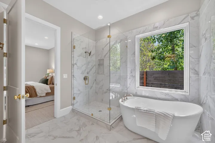 Freestanding soaking tub, walk-in shower with glass enclosure, large window, and marble-look tile flooring