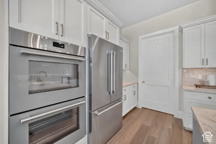 Built-in stainless steel double ovens, stainless steel refrigerator, white shaker-style cabinetry, light wood-finish flooring, and light-toned countertops
