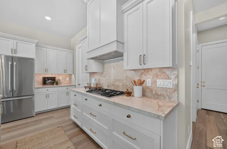White shaker cabinetry with brass-finish hardware, light-toned stone countertops, a coordinating stone backsplash, and stainless steel appliances