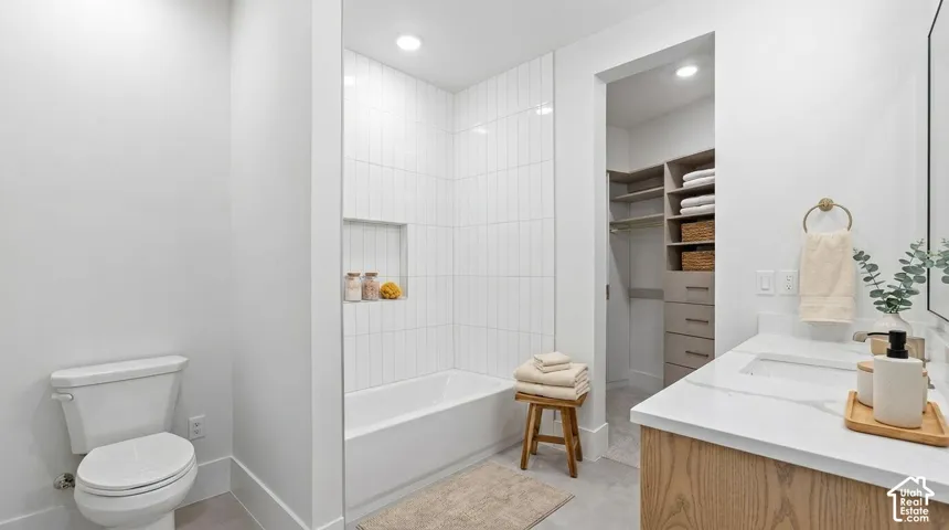 Bathroom featuring a white bathtub with tiled surround and recessed niche, a toilet, a vanity with a white countertop and integrated sink, and an adjacent walk-in closet with built-in shelving and drawers