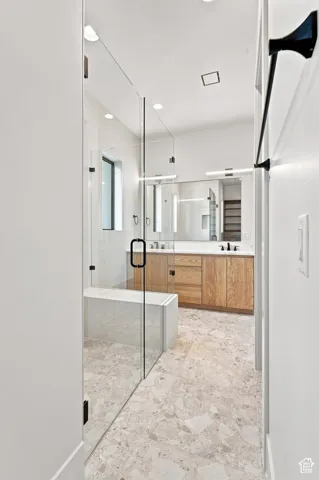 Frameless glass shower enclosure with built-in bench, dual vanities with wood-finish cabinetry, recessed lighting, and stone-finish flooring