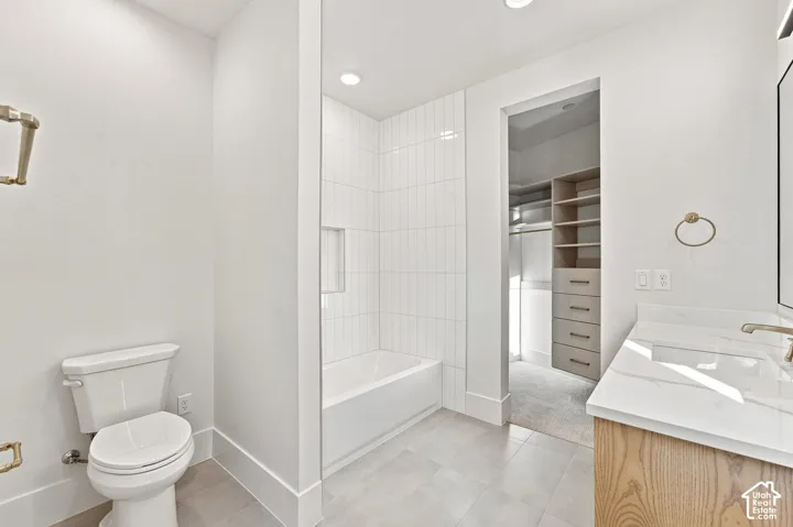 White tile flooring, built-in bathtub with vertical tile surround, wood-finish vanity with countertop and integrated sink, and recessed lighting