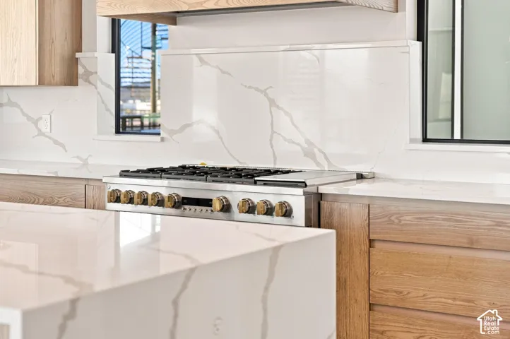 Full height backsplash, under-cabinet windows, stainless steel range, wood-finish cabinetry, and waterfall-edge countertop