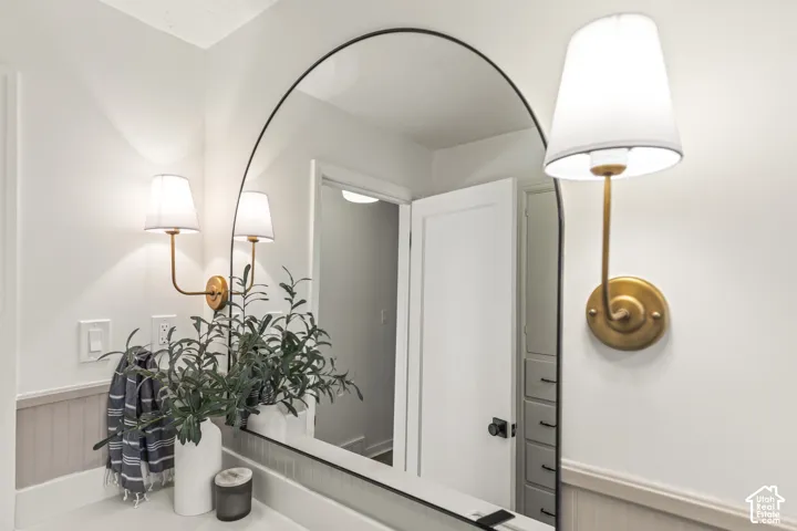 Arch-top vanity mirror with brushed gold wall sconces, beadboard wainscoting, and white countertops
