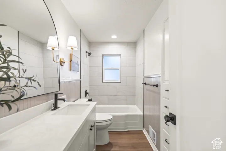 White vanity with matte black hardware and a solid surface countertop, large circular mirror with sconce lighting, white subway tile shower surround, and rich wood-finish flooring
