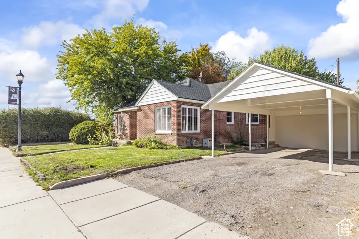 Brick exterior with white trim, covered carport, mature trees, and a concrete sidewalk