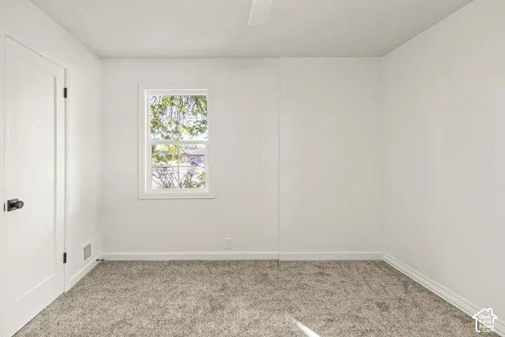 Window with a white frame, light gray wall-to-wall carpeting, white door with dark hardware, and a white ceiling fan