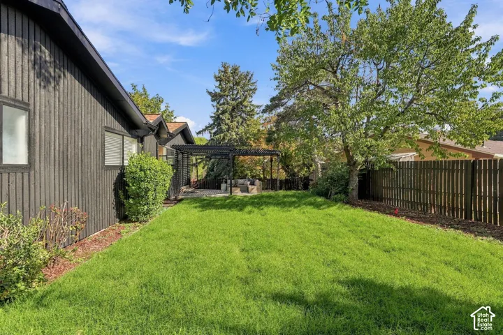 Expansive grassy yard with mature trees, a wooden fence, and a black pergola over a patio area