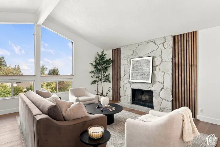 Living area with vaulted ceilings, large picture windows, a stone-clad fireplace, and wood-finish flooring