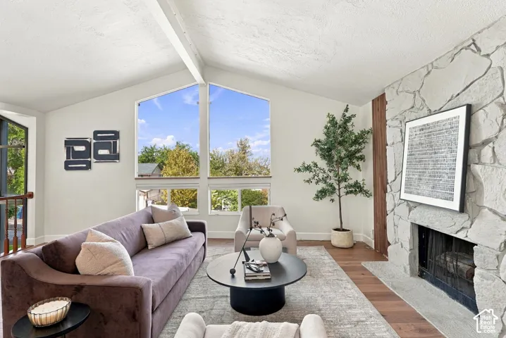 Vaulted ceiling with exposed beams, stone-clad fireplace, large picture windows, wood-finish flooring, and a built-in railing