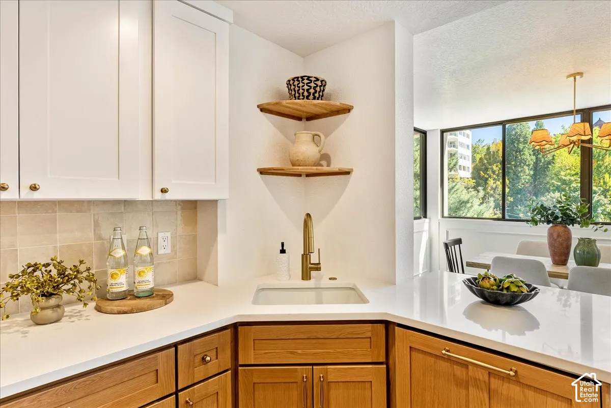 White upper cabinetry with brass hardware, wood-finish lower cabinetry, white solid surface countertops, a single basin sink with a gold-tone faucet, and a stone tile backsplash