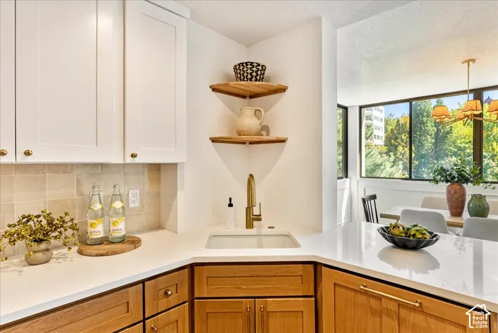 White upper cabinetry with brass hardware, wood-finish lower cabinetry, white solid surface countertops, a single basin sink with a gold-tone faucet, and a stone tile backsplash