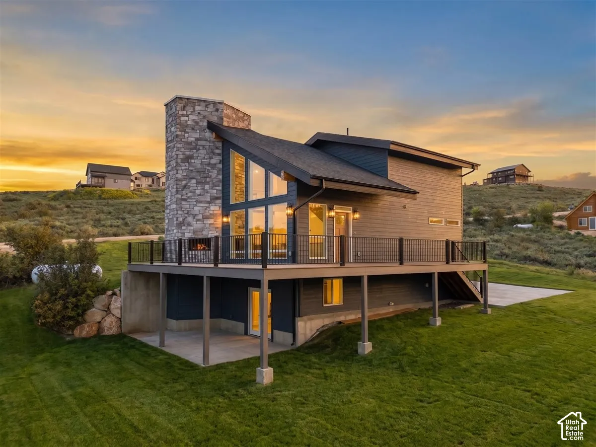 Modern exterior with a stone chimney feature, large deck with dark railings, expansive windows, a covered patio, and a sloped roofline